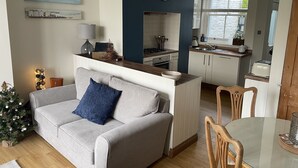Un salon moderne avec un canapé gris, un petit sapin de Noël et un coin cuisine doté de meubles blancs et d’un mur d’accent bleu.