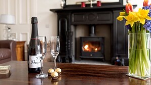 A bottle of Prosecco, two glasses, and a vase with flowers on a wooden table in a room with a fireplace.