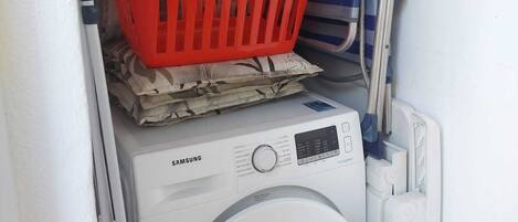 A compact laundry area with a Samsung washing machine, a red plastic basket, and a striped towel rack.