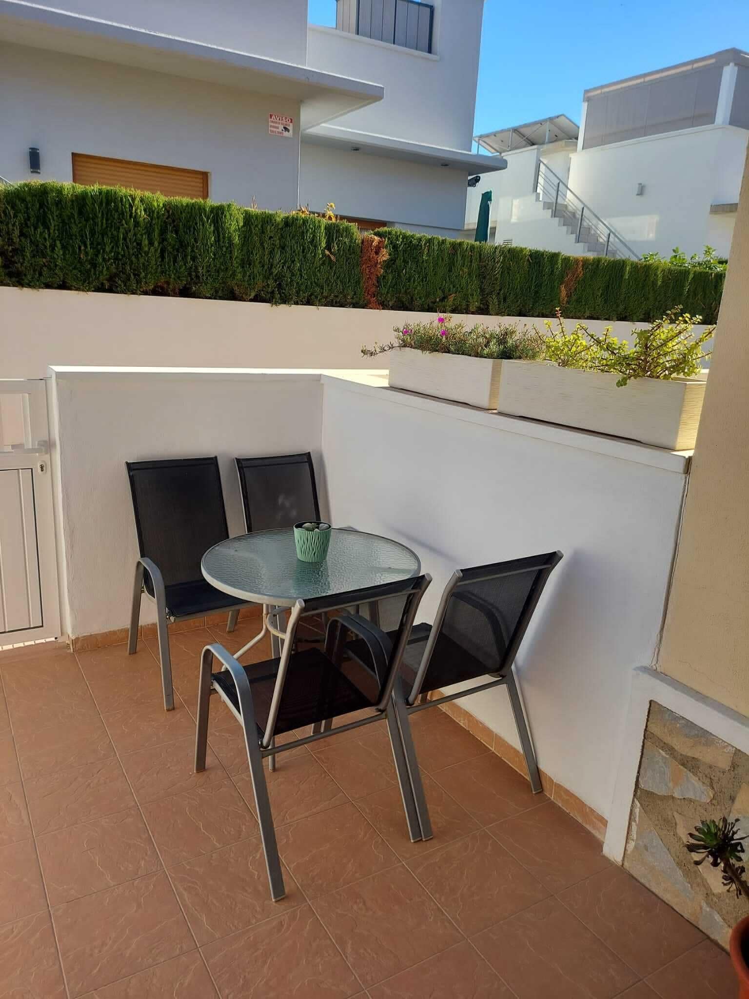 A modern outdoor patio with a glass table and metal chairs, surrounded by white walls and a green hedge.