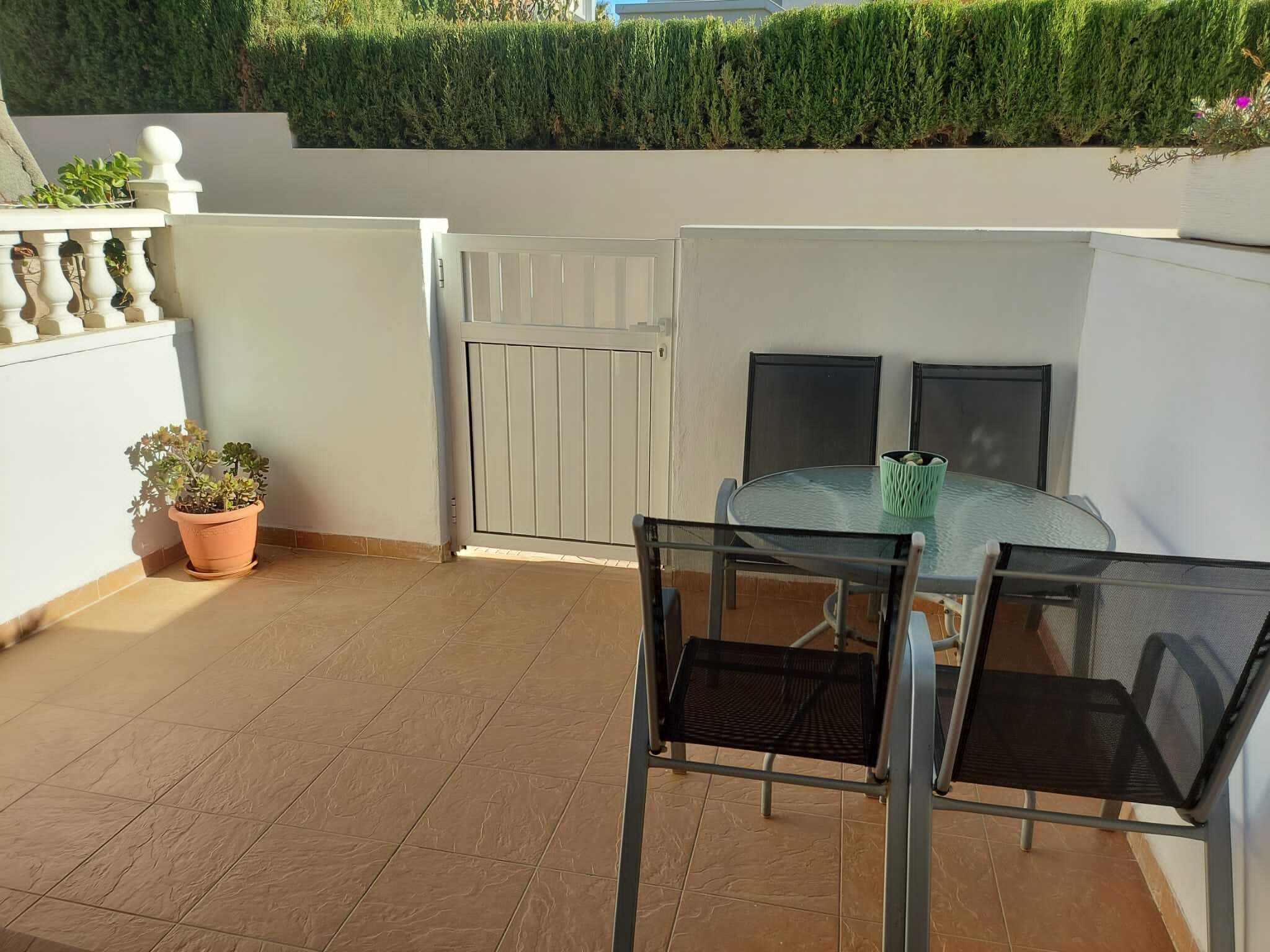 A small outdoor patio with a round glass table and two chairs, a potted plant, and a white gate.