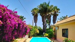 Una piscina rodeada de exuberante vegetación y flores coloridas, con altas palmeras y un edificio con techo de tejas al fondo.