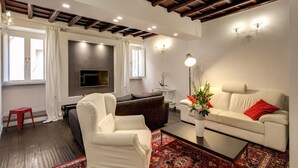 Una sala de estar con un sofá, un sillón y un televisor montado en la pared. Hay una mesita con una lámpara y un jarrón con flores.