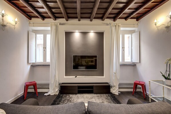 Un salon avec un téléviseur à écran plat fixé au mur, deux tabourets rouges, un canapé gris et un rideau blanc.