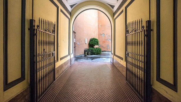 Corredor com piso de azulejos, portões de metal em ambos os lados e uma pequena área de jardim com arbusto e plantas em vasos.