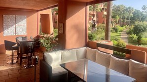 A sunlit room with a glass table, white cushioned seating, and a view of a landscaped garden. - Golf, Tennis, Paddle Tennis, Gym, 4 pools including heated, (Ojen)