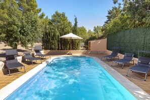 Et rektangulært swimmingpool med flisekant, omgivet af liggestole og en pergola med parasol.