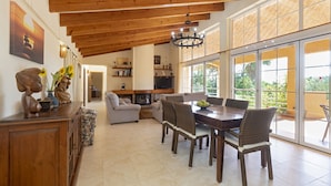Amplia sala de estar con techo alto, vigas de madera, chimenea, mesa de comedor con sillas y ventanas grandes que ofrecen vista a la vegetación.