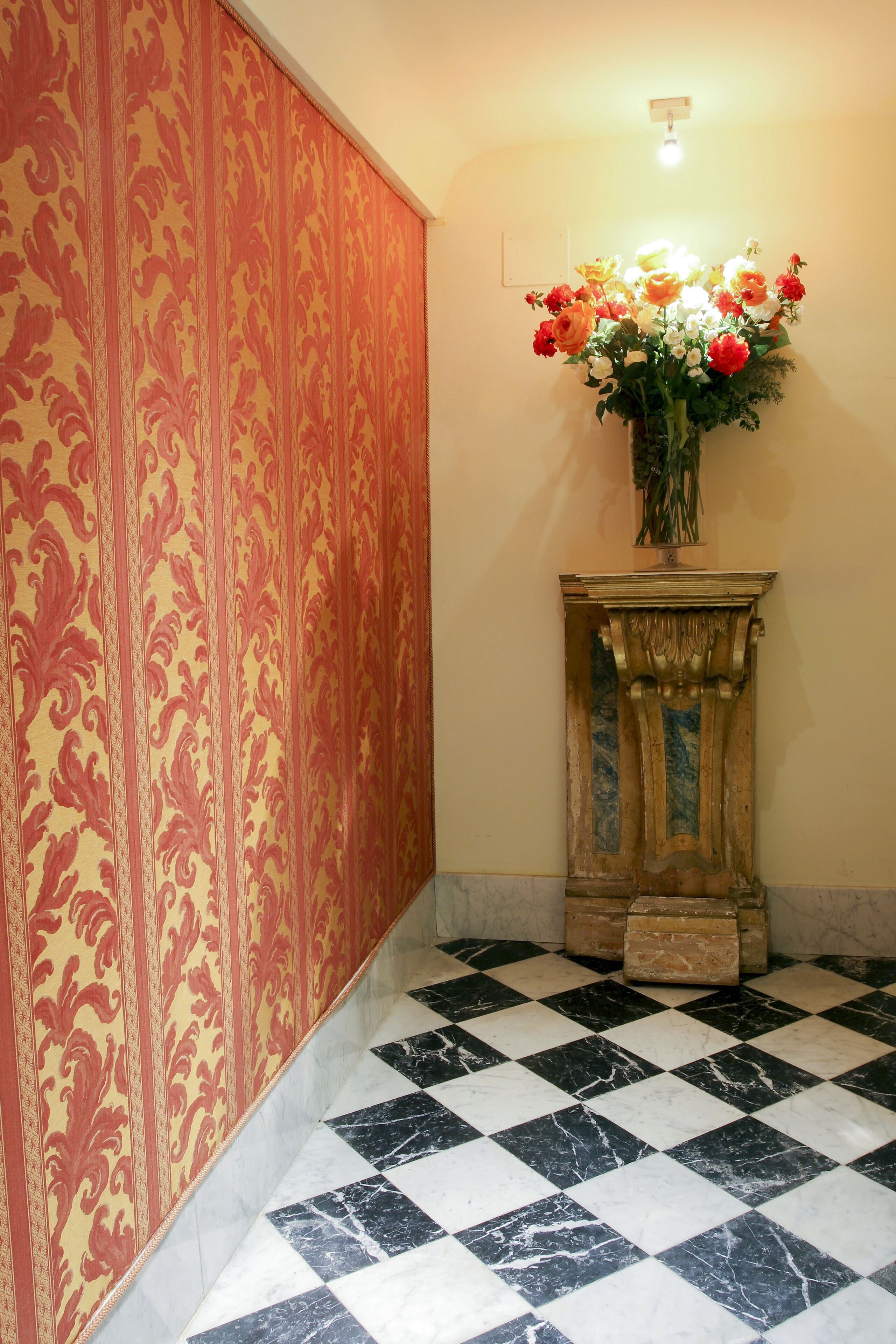 A checkered floor, a patterned wall, and a vase with flowers on a pedestal.