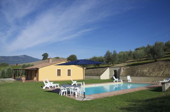 A pool area with white lounge chairs, a blue umbrella, and a yellow building.