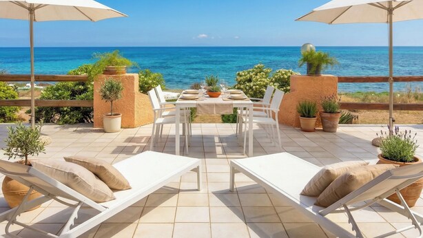 Una terraza con una mesa para dos, sillas de lounge blancas, plantas en macetas y vistas al mar.