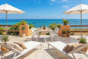 Una terraza con una mesa para dos, sillas de lounge blancas, plantas en macetas y vistas al mar.