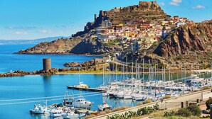 Un pueblo costero con una fortaleza histórica en una colina, un puerto deportivo con barcos, y una carretera que lleva al pueblo.