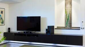 Een moderne woonkamer met een flatscreen televisie op een zwarte televisiemeubel, een decoratieve plant in een raam en een kunstwerk in een lijst aan de muur.