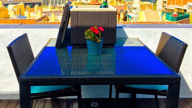 Une terrasse sur le toit, dotée d’une table en verre et de chaises en osier, offrant une vue sur un paysage urbain avec des bâtiments et une colline au loin.