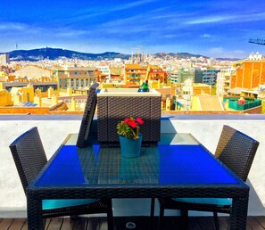 A rooftop terrace with a glass table and wicker chairs, overlooking a cityscape with buildings and a hill in the distance. - BEAUTIFUL PANORAMIC PENTHOUSE + 20m2 TERRACE at 800m BEACH/BORN/GÓTIC QUARTERS (Barcelona)