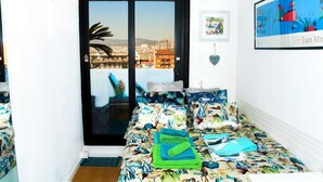 Une petite pièce, bien éclairée, avec vue sur la ville à travers une porte coulissante. On y trouve un lit recouvert d’une courtepointe à motifs floraux, un tableau encadré au mur et une petite étagère avec une plante.