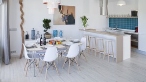 Ett modernt kök med en central köksö, vita barstolar och en matplats med vita stolar och ett bord som är dukat för fyra personer.