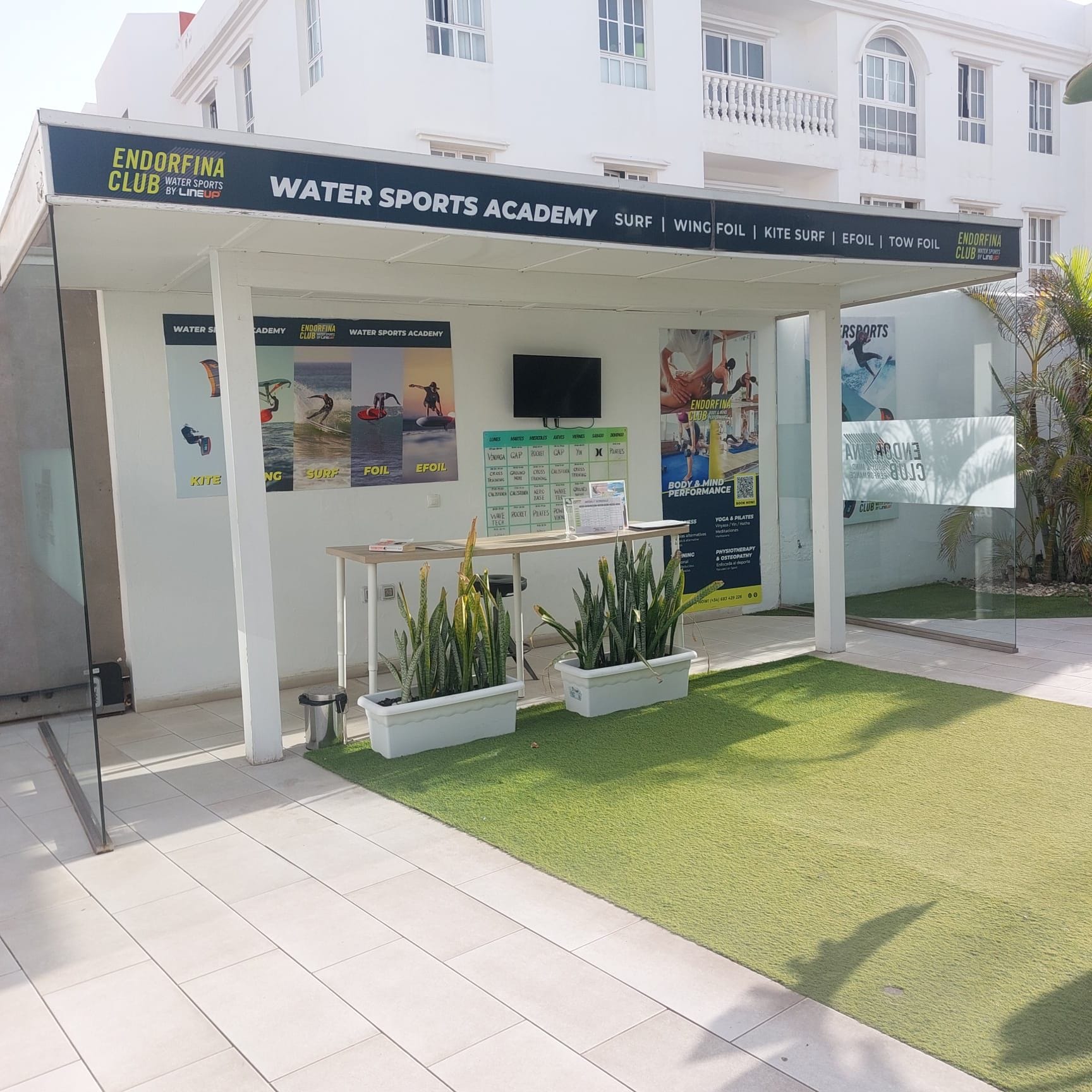 Een watersportacademie met een overdekt zitgedeelte, posters van watersportactiviteiten en een kleine tafel met planten.