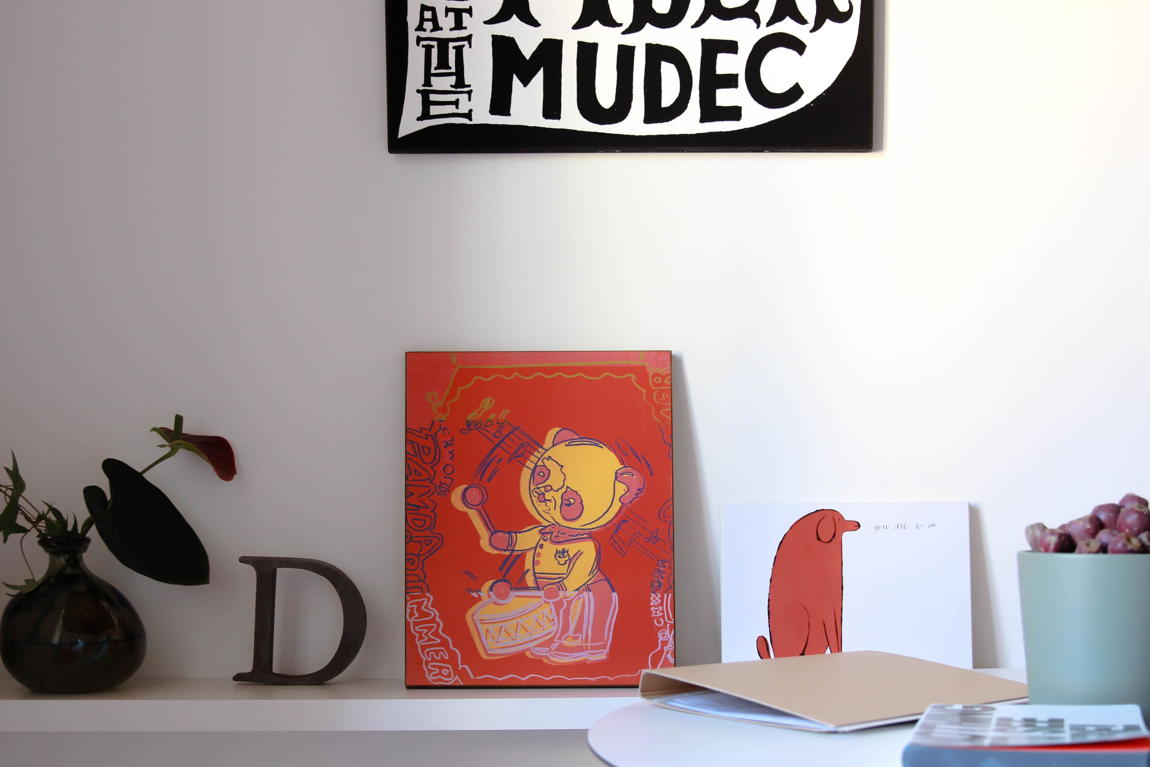 Un mur avec un panneau encadré, une étagère avec un vase, la lettre « D », une peinture colorée et une carte avec un dessin.