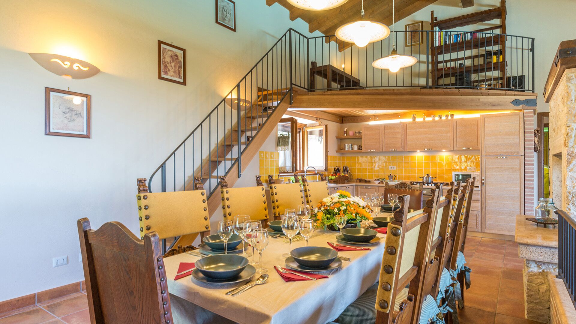 Un comedor con una larga mesa dispuesta para una comida, una escalera que sube a un nivel superior y una cocina al fondo.