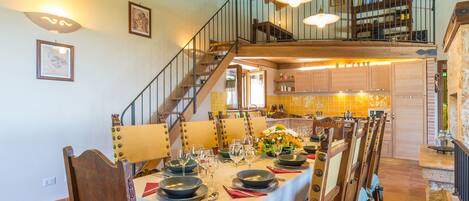 Un comedor con una larga mesa dispuesta para una comida, una escalera que sube a un nivel superior y una cocina al fondo.