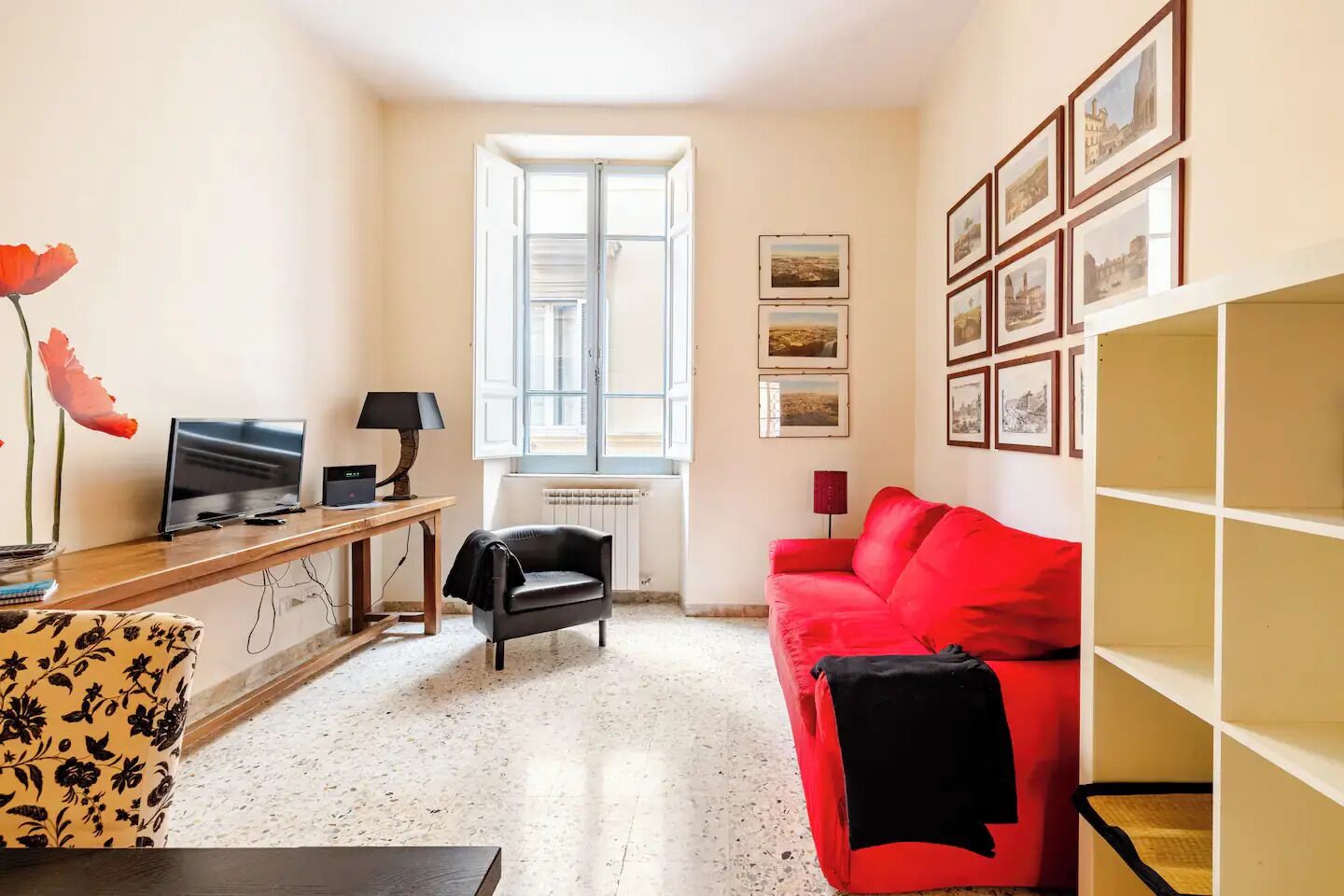 Un salon avec un téléviseur, un bureau, un canapé rouge et des tableaux encadrés accrochés au mur.