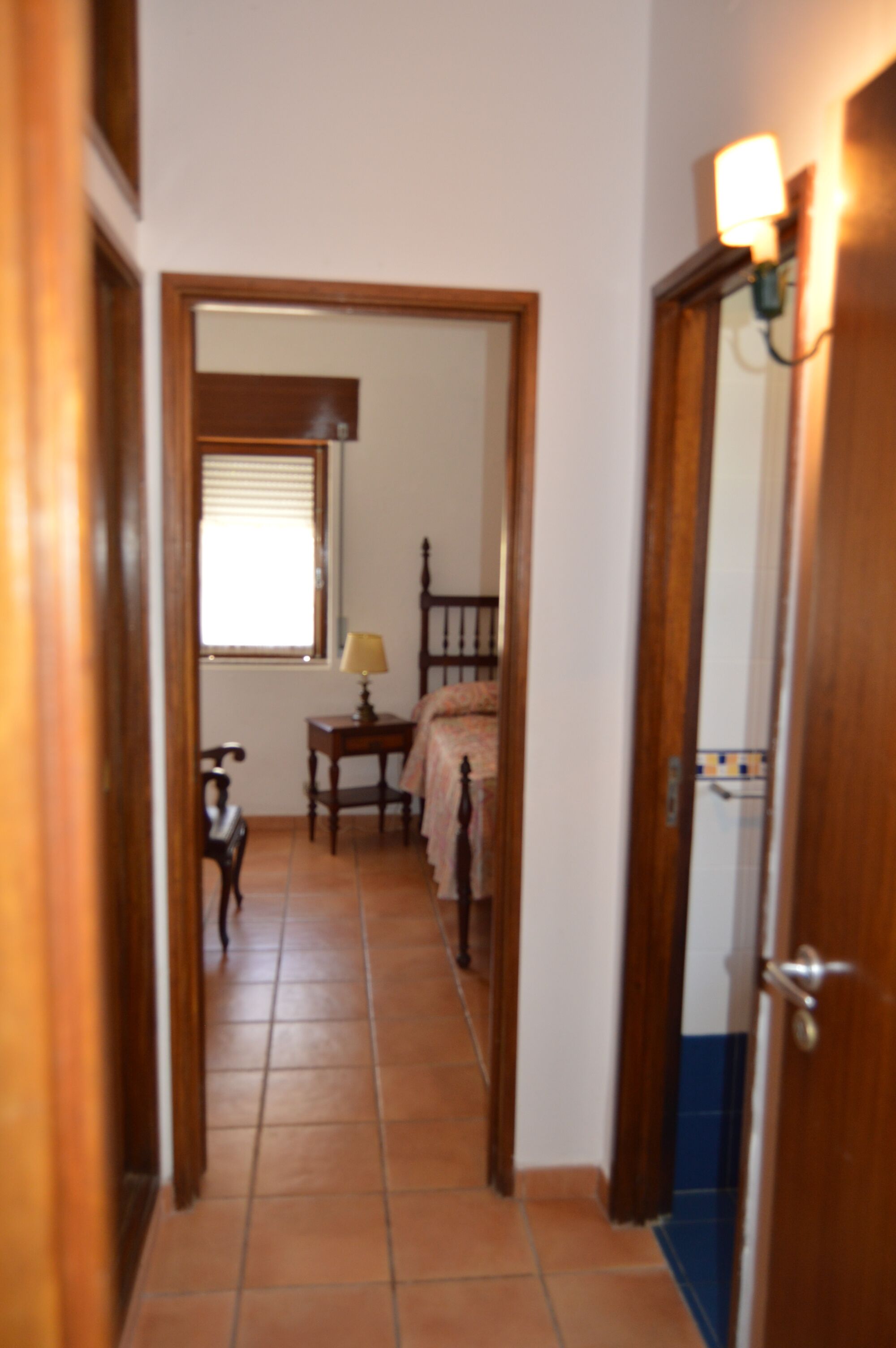 Un couloir étroit avec une porte en bois sur la droite, une fenêtre avec des stores sur la gauche, et un lit avec une table de chevet à l’arrière-plan.