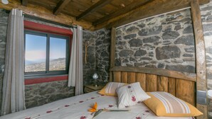 Een slaapkamer met stenen muren, een houten plafond, een bed met bloemenbeddengoed en een raam met gordijnen.