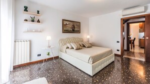 Un dormitorio con cama, mesitas de noche, radiador, estante con objetos decorativos, un cuadro y una puerta que da a otra habitación.