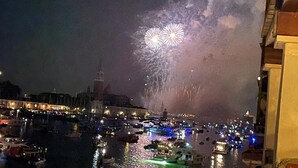 Una vista nocturna de un puerto con barcos y edificios, y fuegos artificiales en el cielo.