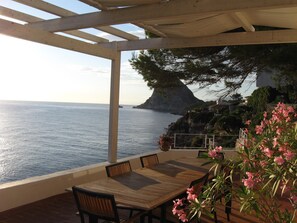 A wooden table and chairs under a pergola with a view of the sea and a rocky coastline.