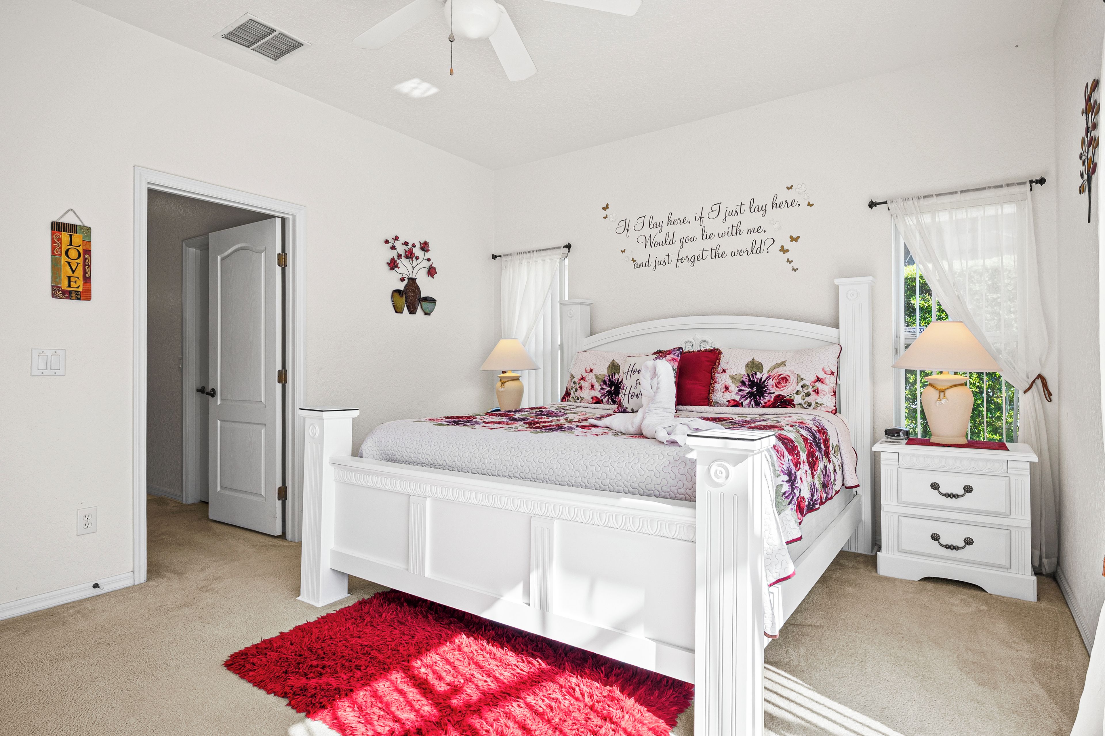 A bedroom with a white bed, a red rug, a wall clock, and a window with curtains.