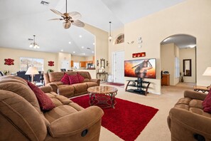 A spacious living room with a large flat-screen TV, comfortable sofas, a red rug, and a ceiling fan.