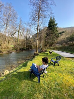 Une personne assise sur une chaise, au bord d’une rivière, est en train de lire un livre.