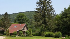 A house with a sloped roof and a small attached garage, surrounded by a well-maintained lawn and dense forest. - Very beautiful 3* Cottage on the riverside, quiet in a green setting (Nans-Sous-Sainte-Anne)