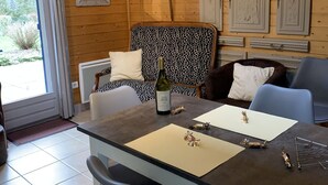 A cozy room with a wooden interior, a dining table with chairs, a bottle of wine, decorative plates on the wall, and a view of the outdoors. - Very beautiful 3* Cottage on the riverside, quiet in a green setting (Nans-Sous-Sainte-Anne)