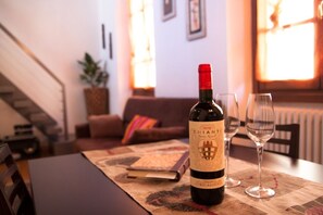 A bottle of Chianti wine on a table with a glass, in a living room with a staircase, a plant, and a window.