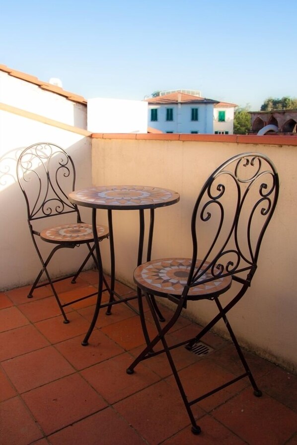 A small outdoor patio with two metal chairs and a round table, featuring a tiled floor and a plain wall in the background.
