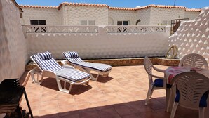 A patio with two sun loungers, a table with chairs, and a grill.