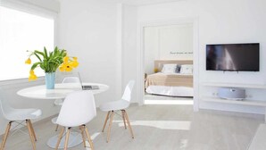 A modern hotel room with a round dining table, white chairs, a potted plant, a bed with a headboard, and a mounted TV. - Sitges Bellavista Spectacular Sea Views (Sitges)