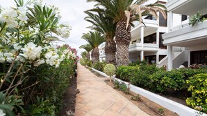 Un chemin bordé d’une végétation luxuriante et de fleurs blanches, qui mène à un bâtiment moderne doté de nombreux balcons et d’une façade blanche.