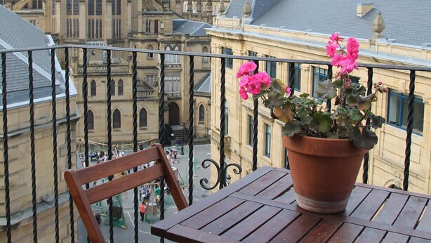 A balcony with a wooden table and chair, a potted plant with flowers, and a view of a historic building with a spire. - Apartment overlooking the cathedral, terrace, wifi, parking included (Donostia)