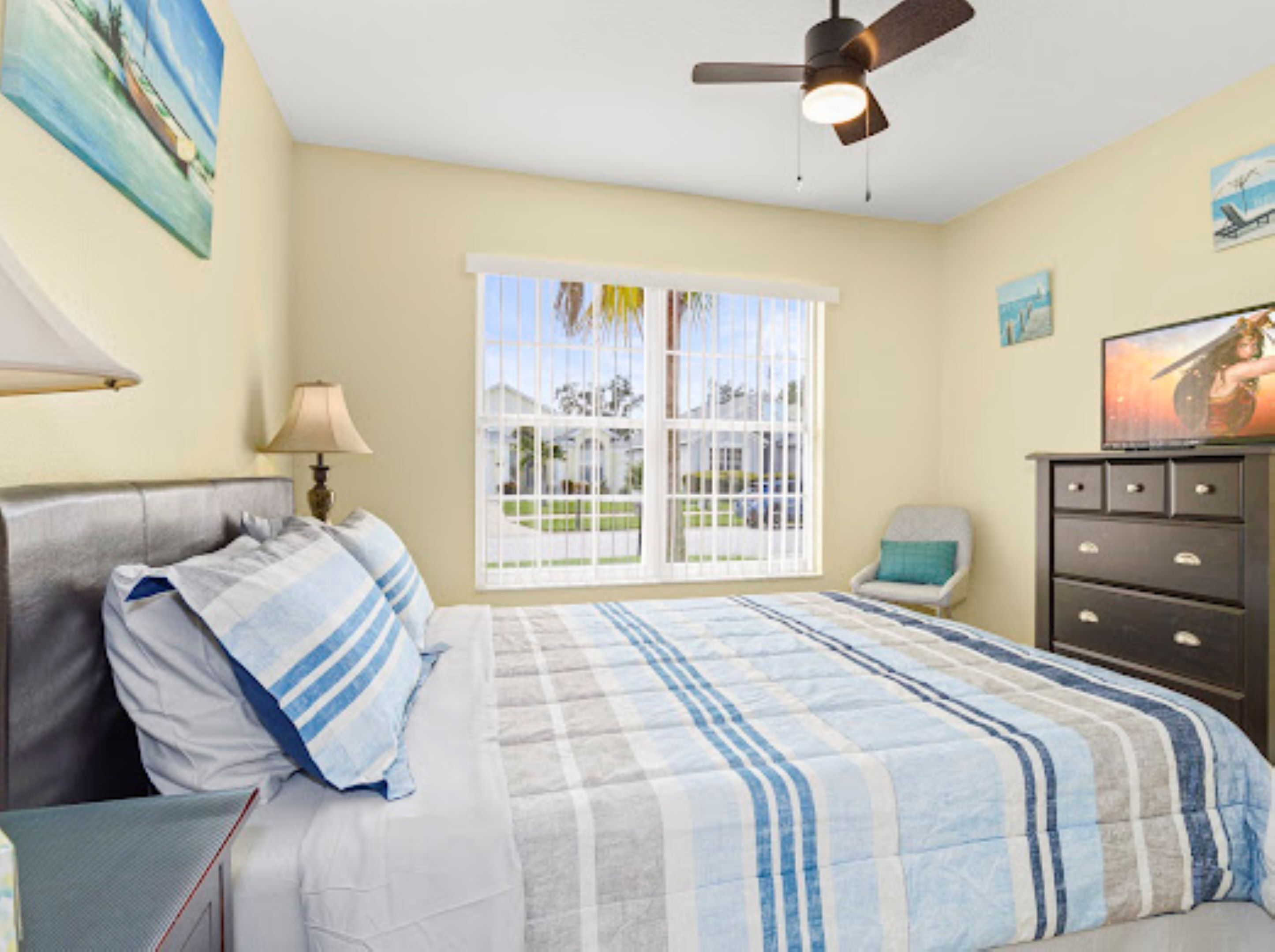 A bedroom with a bed, a ceiling fan, a window with a view, a chair, and a dresser.
