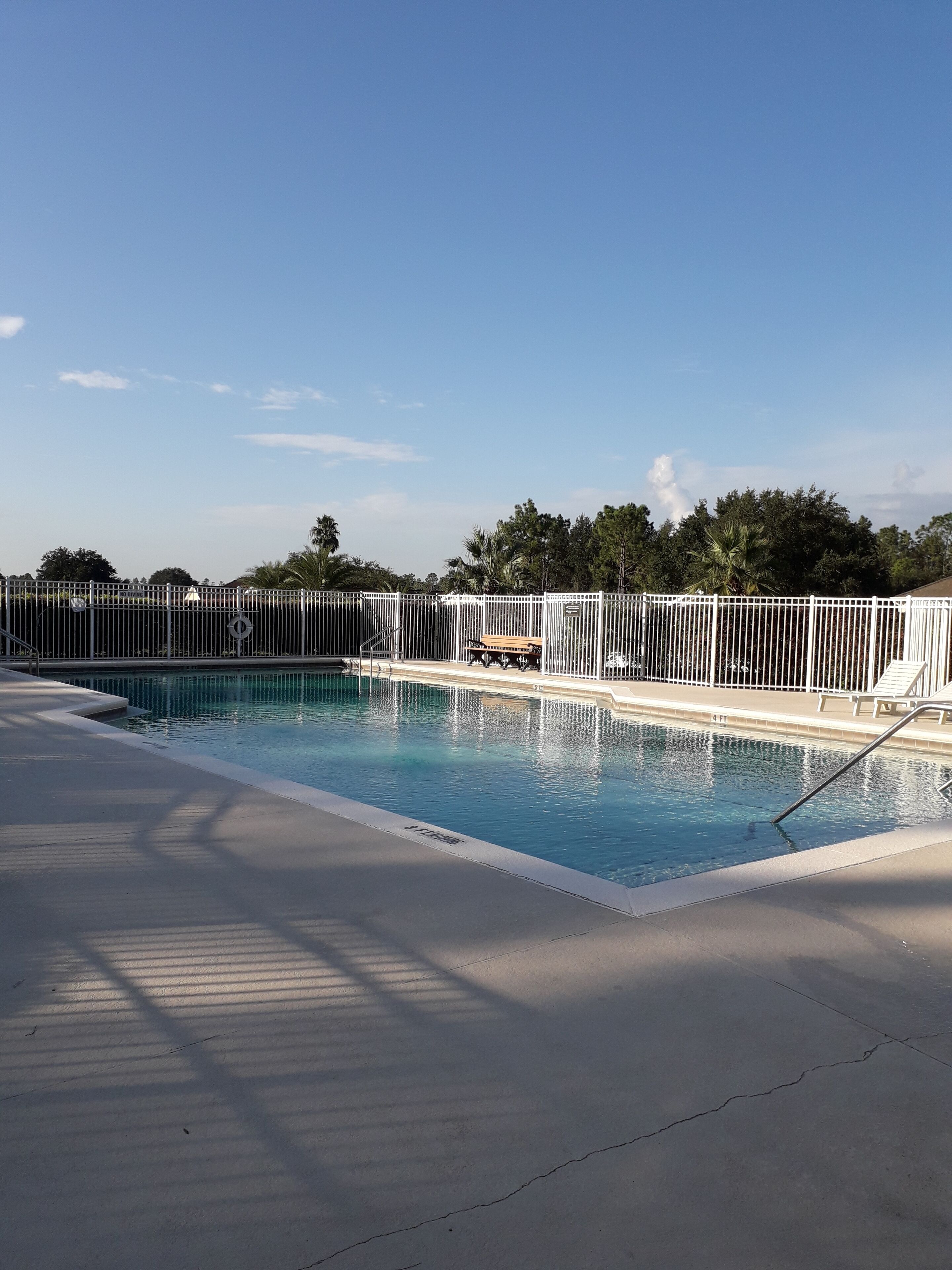 Una piscina con valla blanca, una banca y una zona con sombra.
