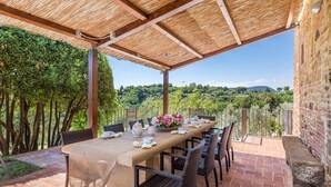 An outdoor patio with a table set for dining, surrounded by wicker chairs, under a thatched roof, with a view of greenery and hills in the background. - Bossoncina 8+1, Emma Villas (Montepulciano)