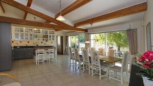 Una cuina àmplia amb armaris blancs, zona de dinar amb taula de fusta i cadires blanques, i grans finestres que donen vistes a verdura exterior.