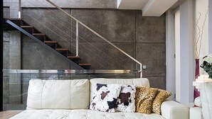 Una sala de estar moderna con una mesa de centro de vidrio, un sofá con cojines estampados y una escalera con barandilla metálica.