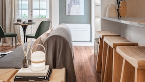 Una habitación de hotel moderna con una cama grande, un comedor con mesa y sillas, una cocina con taburetes de madera, y vistas al exterior a través de un amplio ventanal.