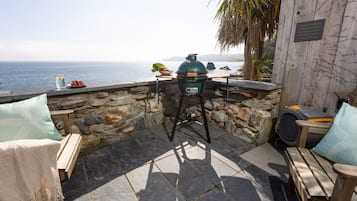 A stone-walled outdoor seating area with a view of the sea, a grill, and a wooden table with a bowl of fruit.
